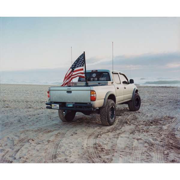 White pickup truck on a beach with an American flag waving from the truck bed, showcasing a coastal scene with the ocean in the background.