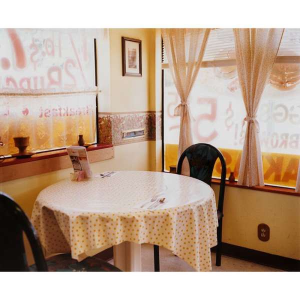 Diner interior with round table, polka dot tablecloth, and curtained windows showing exterior restaurant signage in soft, diffused light.