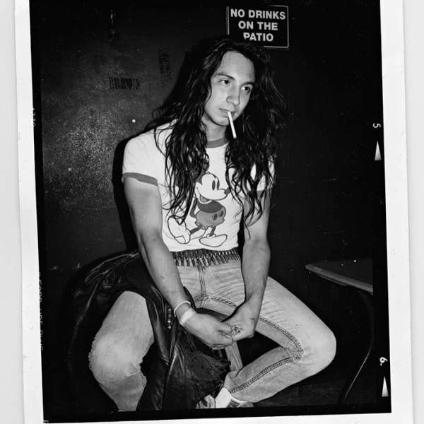 Black and white image of a young adult male with long hair sitting, smoking a cigarette in an indoor setting with a dark background.