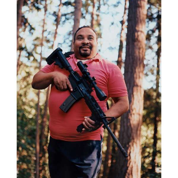Man in pink polo shirt standing in a forest holding a black rifle with an optic sight. Trees provide a blurred background.