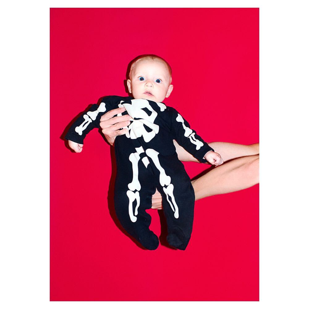 Infant in skeleton costume held in arms against bright red background. Full shot showing the baby and arms holding the baby.