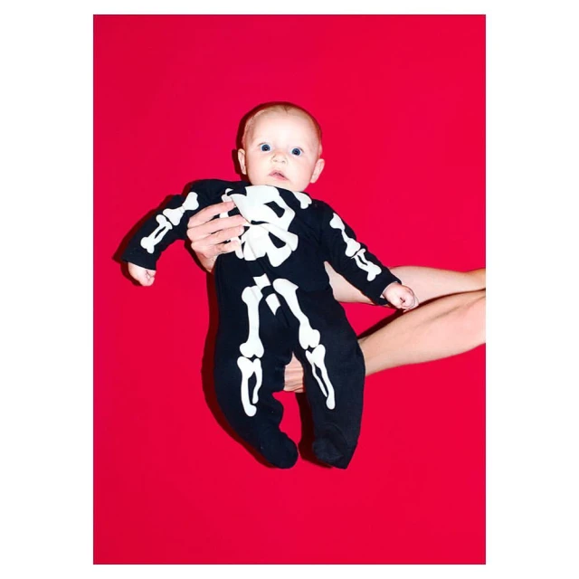 Infant in skeleton costume held in arms against bright red background. Full shot showing the baby and arms holding the baby.