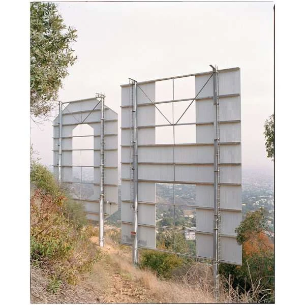 Rear view of the Hollywood sign on a hillside, partially obscured by greenery. The sign is white with a metal framework.