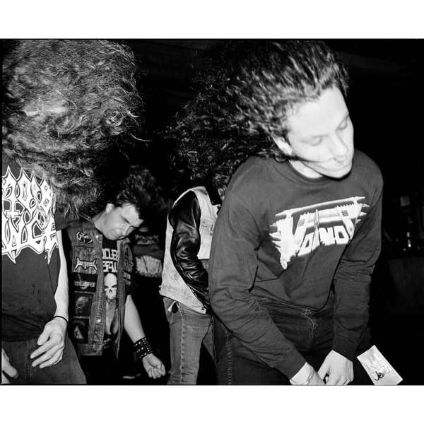 Black and white photo of a group of people with long hair, some wearing vests and band t-shirts, in a dark, indoor setting.