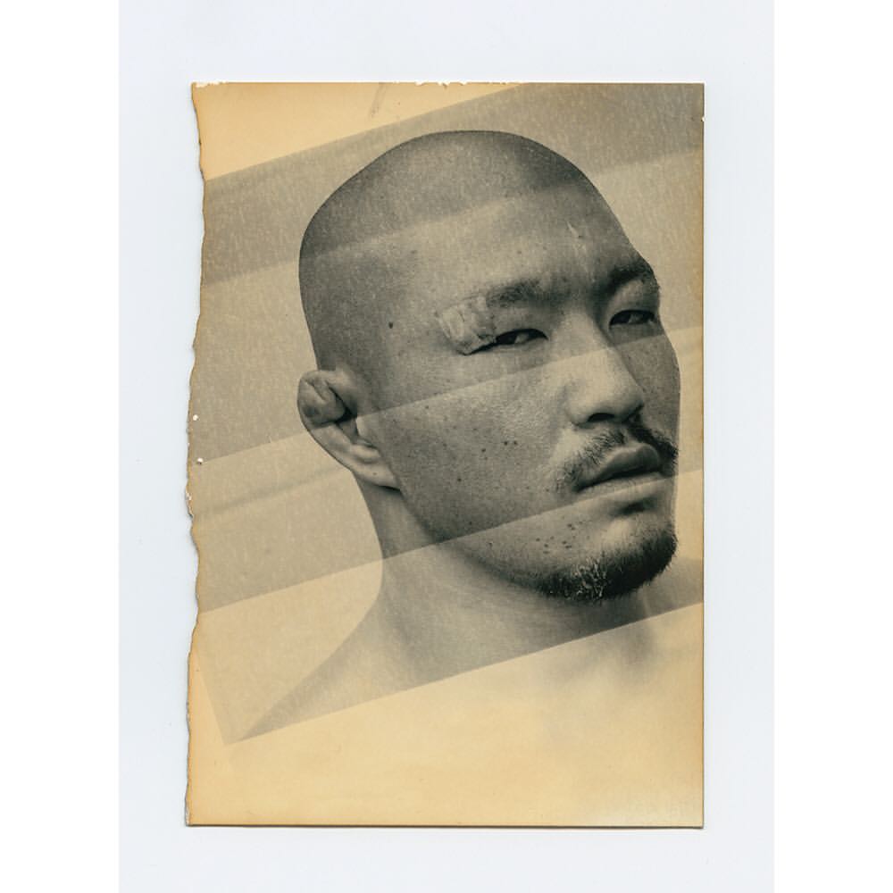 Close-up of a man with a shaved head and a beard in a studio setting. The image is captured on vintage paper with a monochromatic palette.
