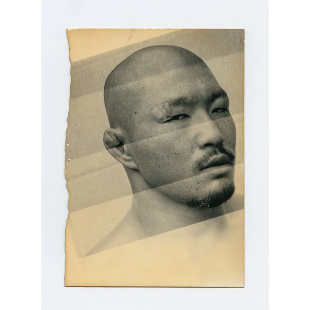 Close-up of a man with a shaved head and a beard in a studio setting. The image is captured on vintage paper with a monochromatic palette.
