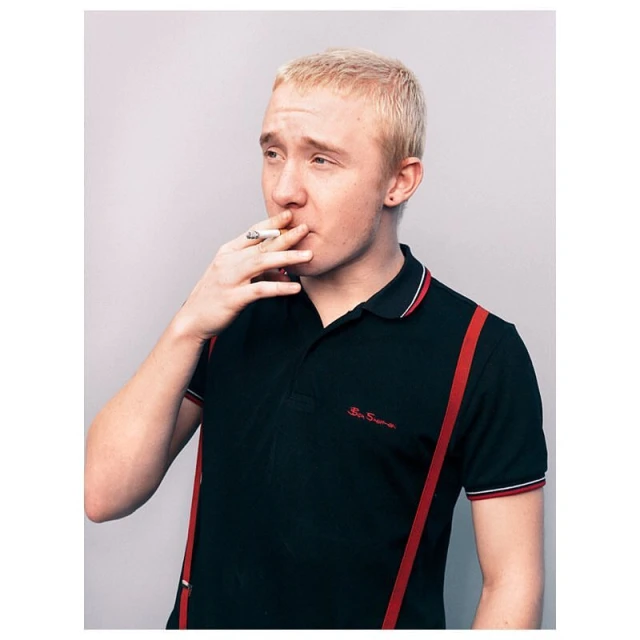 A blond male is smoking a cigarette while wearing a black polo shirt with red suspenders and standing against a grey background.