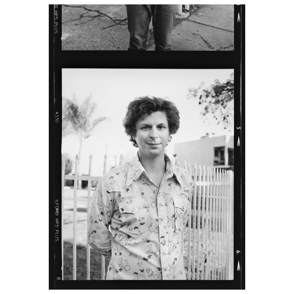 Black and white portrait of adult man in patterned shirt standing in front of white picket fence with building and tree behind.