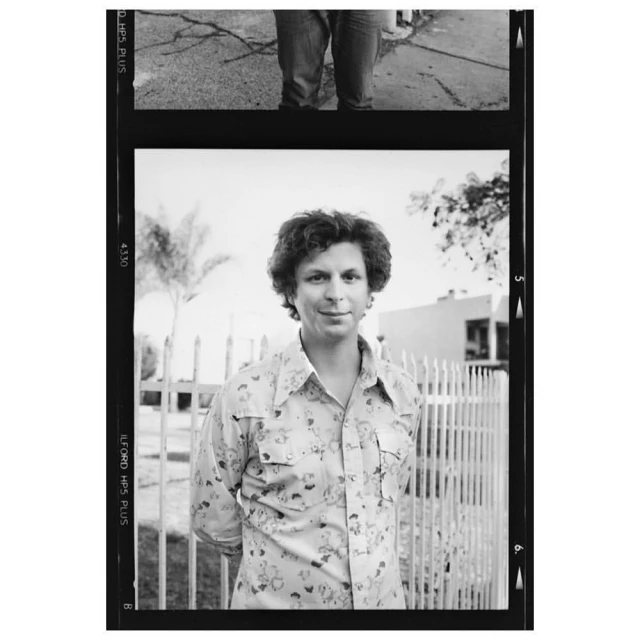 Black and white portrait of adult man in patterned shirt standing in front of white picket fence with building and tree behind.