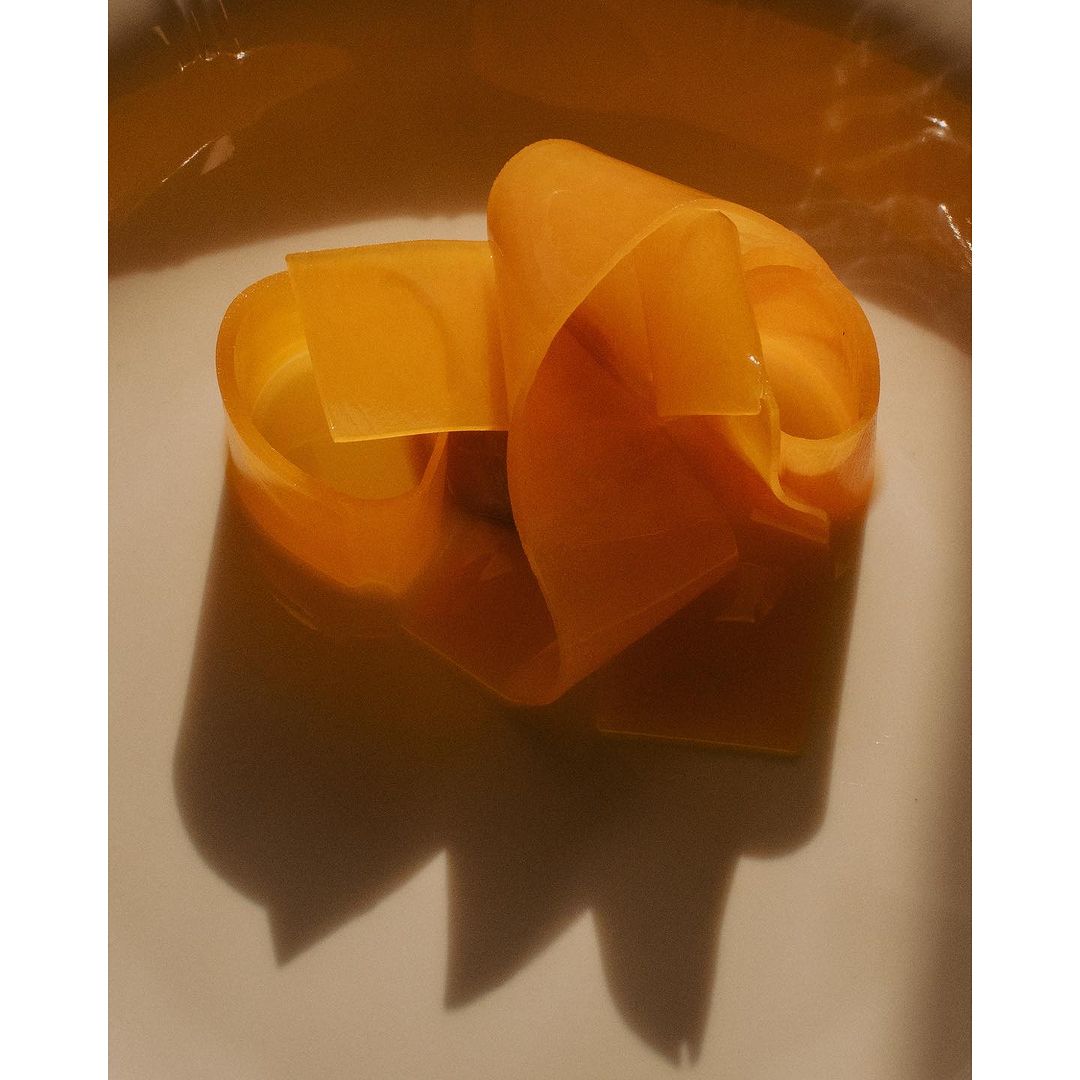 Abstract still life of translucent orange plastic ribbon arranged in sculptural curls on a white surface, casting shadows.