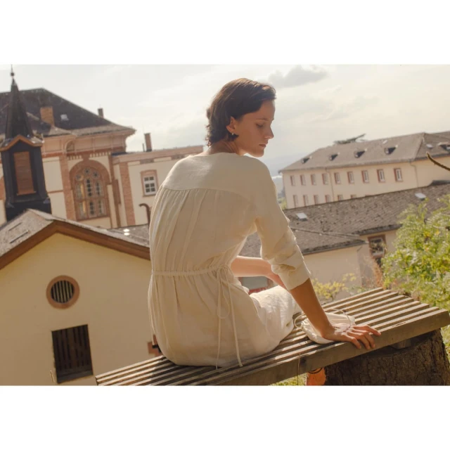 Woman in a cream-colored dress sits on a weathered wooden bench with buildings visible on a sunny day. Editorial fashion lifestyle shoot.