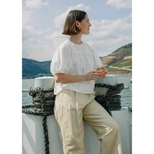 Adult woman in white blouse and neutral pants stands next to the river with hills on the opposite bank under the cloudy sky.