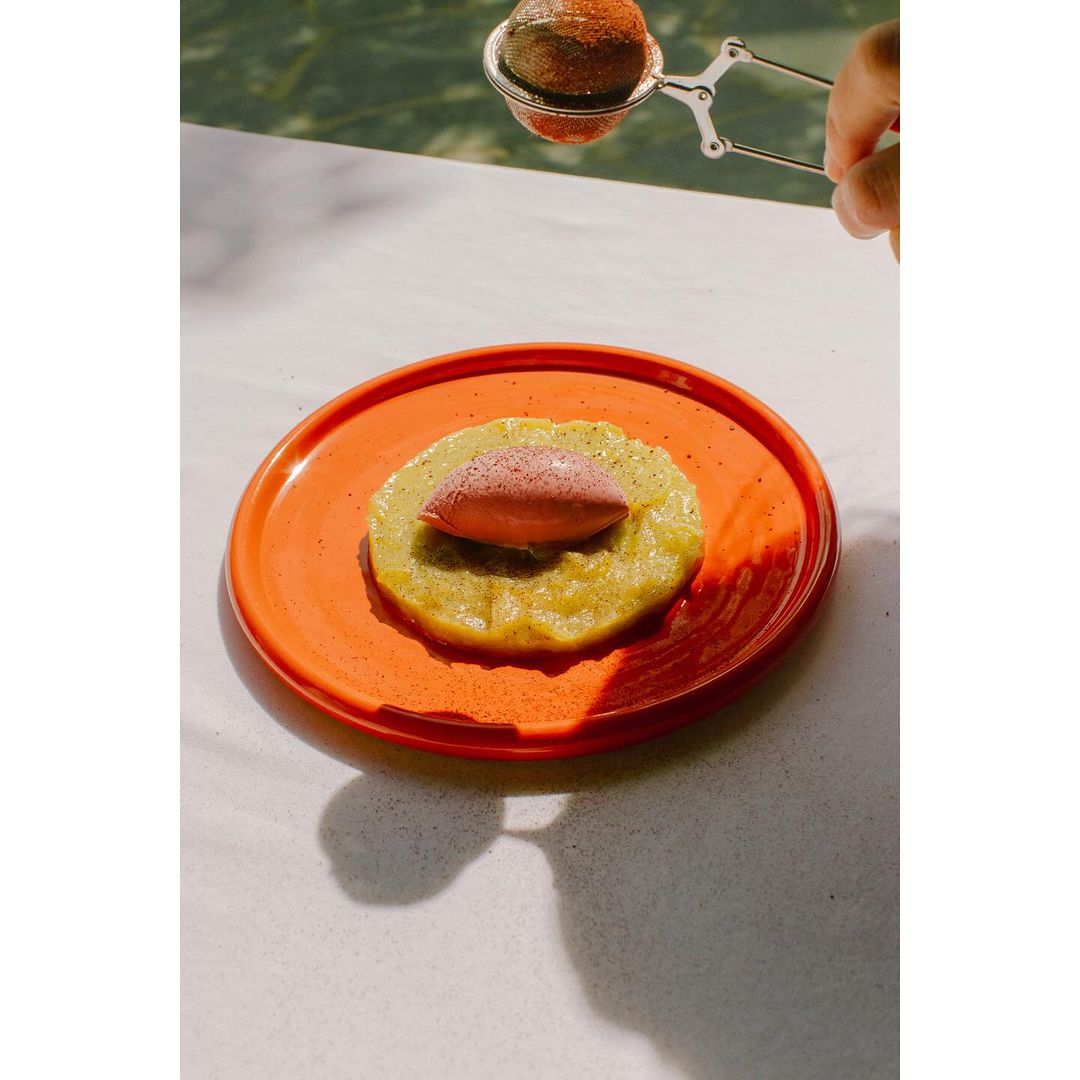 Orange plate topped with dessert and dusted with powder. Hand holds sifter above food. Minimalist food presentation.