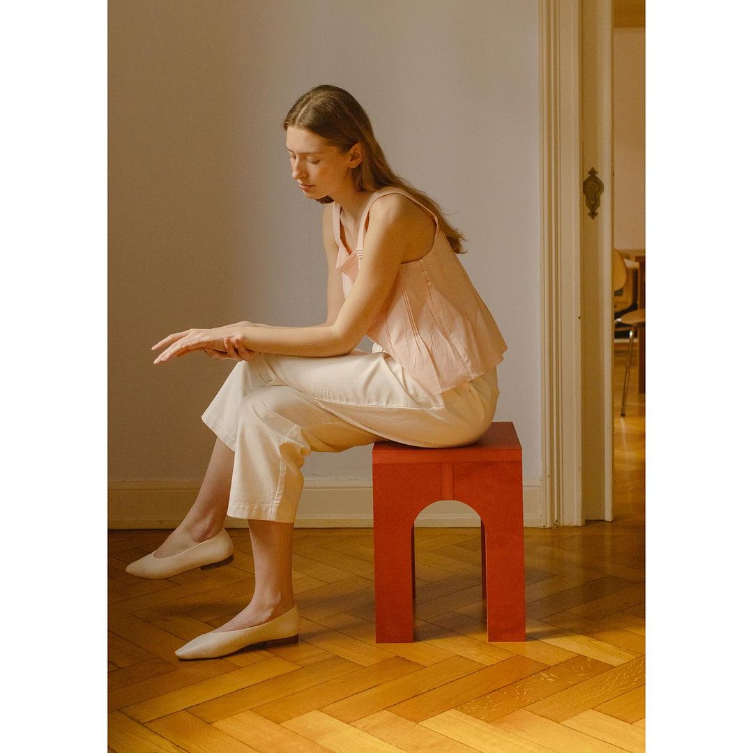 A young woman sits on a red stool in an indoor setting. She is wearing white pants, ballet shoes, and a pink top, creating a minimalist, fashion-forward look.