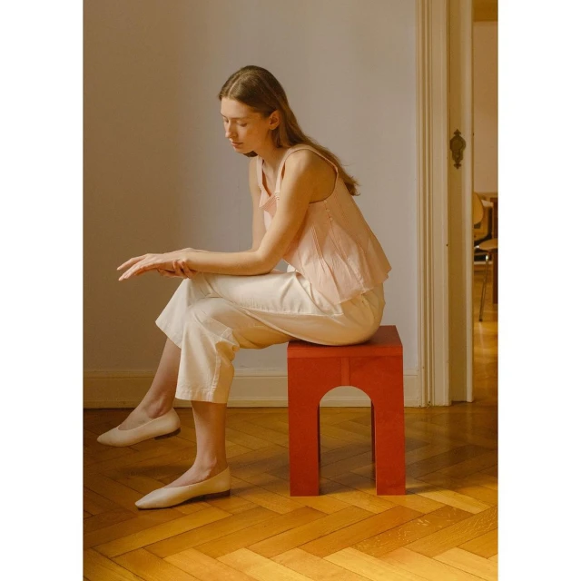 A young woman sits on a red stool in an indoor setting. She is wearing white pants, ballet shoes, and a pink top, creating a minimalist, fashion-forward look.