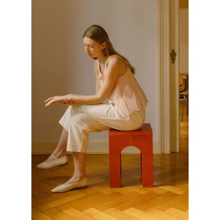 A young woman sits on a red stool in an indoor setting. She is wearing white pants, ballet shoes, and a pink top, creating a minimalist, fashion-forward look.