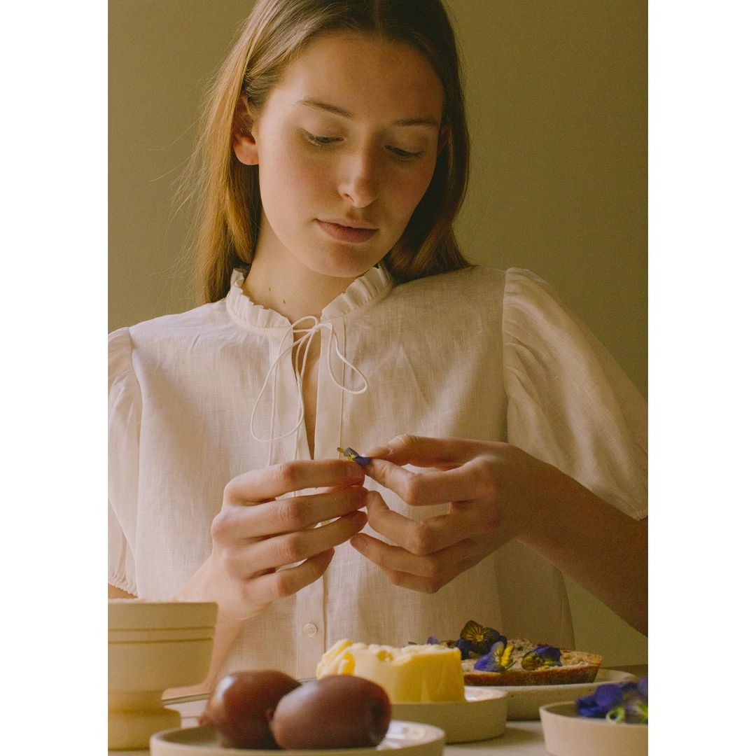 Young woman in a white blouse sits at a table with food, including kiwi and a butter pat topped with blue edible flowers.