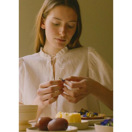 Young woman in a white blouse sits at a table with food, including kiwi and a butter pat topped with blue edible flowers.