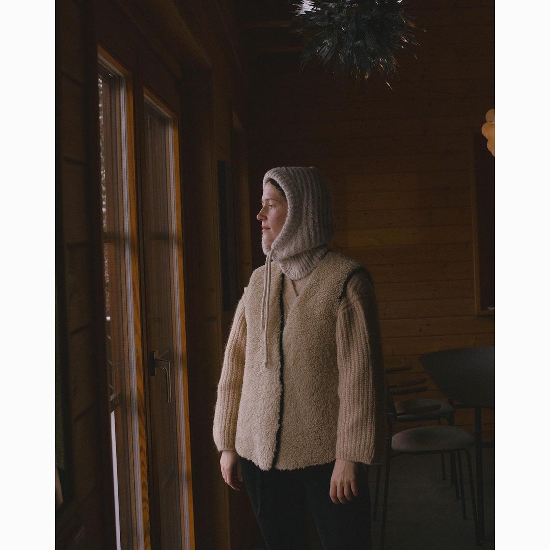 Woman in beige hooded sweater and vest looks out window in wood paneled room, capturing indoor fashion and contemplative mood.