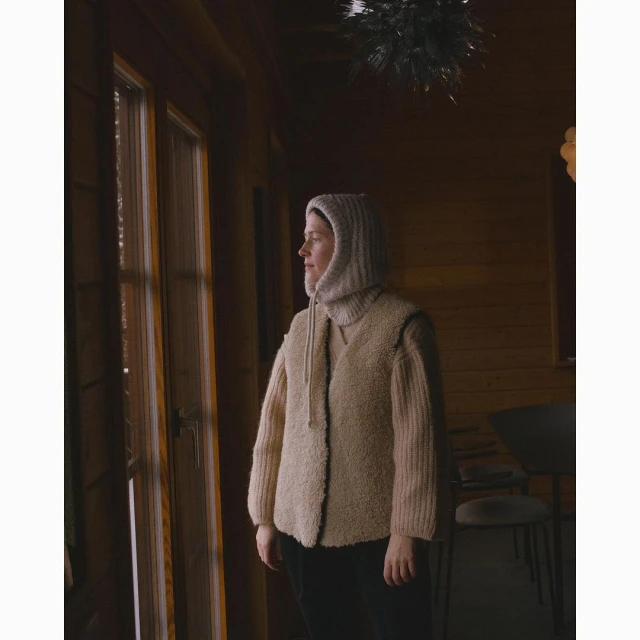 Woman in beige hooded sweater and vest looks out window in wood paneled room, capturing indoor fashion and contemplative mood.