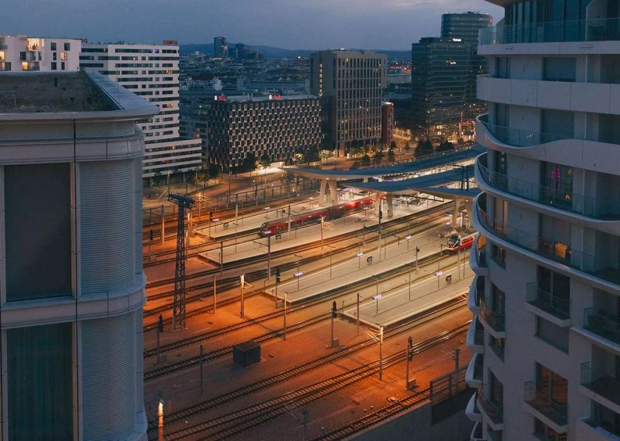 Elevated view of a modern urban railway station with multiple tracks and platforms, surrounded by contemporary buildings at twilight.