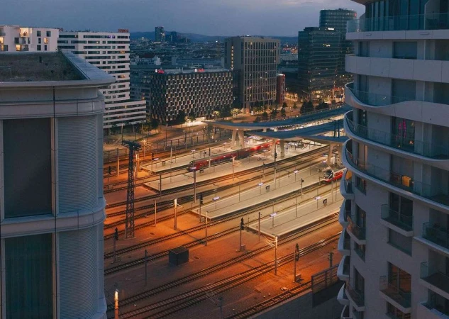 Elevated view of a modern urban railway station with multiple tracks and platforms, surrounded by contemporary buildings at twilight.