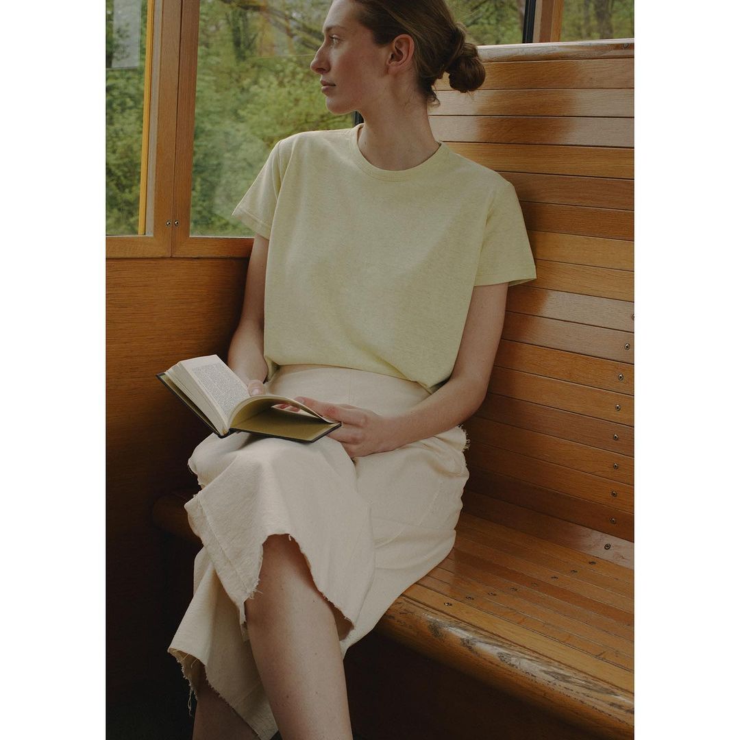 Young woman in casual wear sits on a wooden bench and reads a book by a window. Neutral color palette. Natural light.