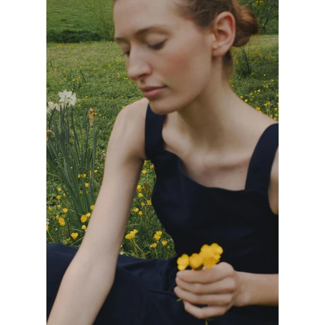 Young woman in dark dress sits in a field of yellow wildflowers, holding a bunch of buttercups in her hand. Nature backdrop.