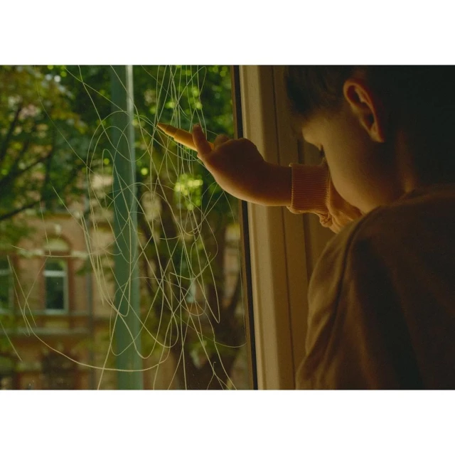 Child drawing on window with crayon. Small child creating art on window with trees and buildings in background on a sunny day.