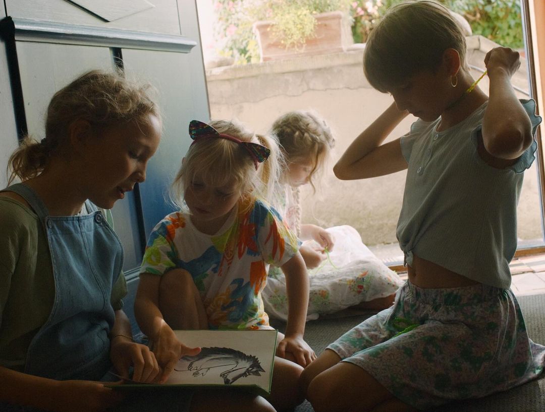 Four young girls gather indoors, one reading aloud from a book with a wolf drawing, casual clothing, and soft natural light.