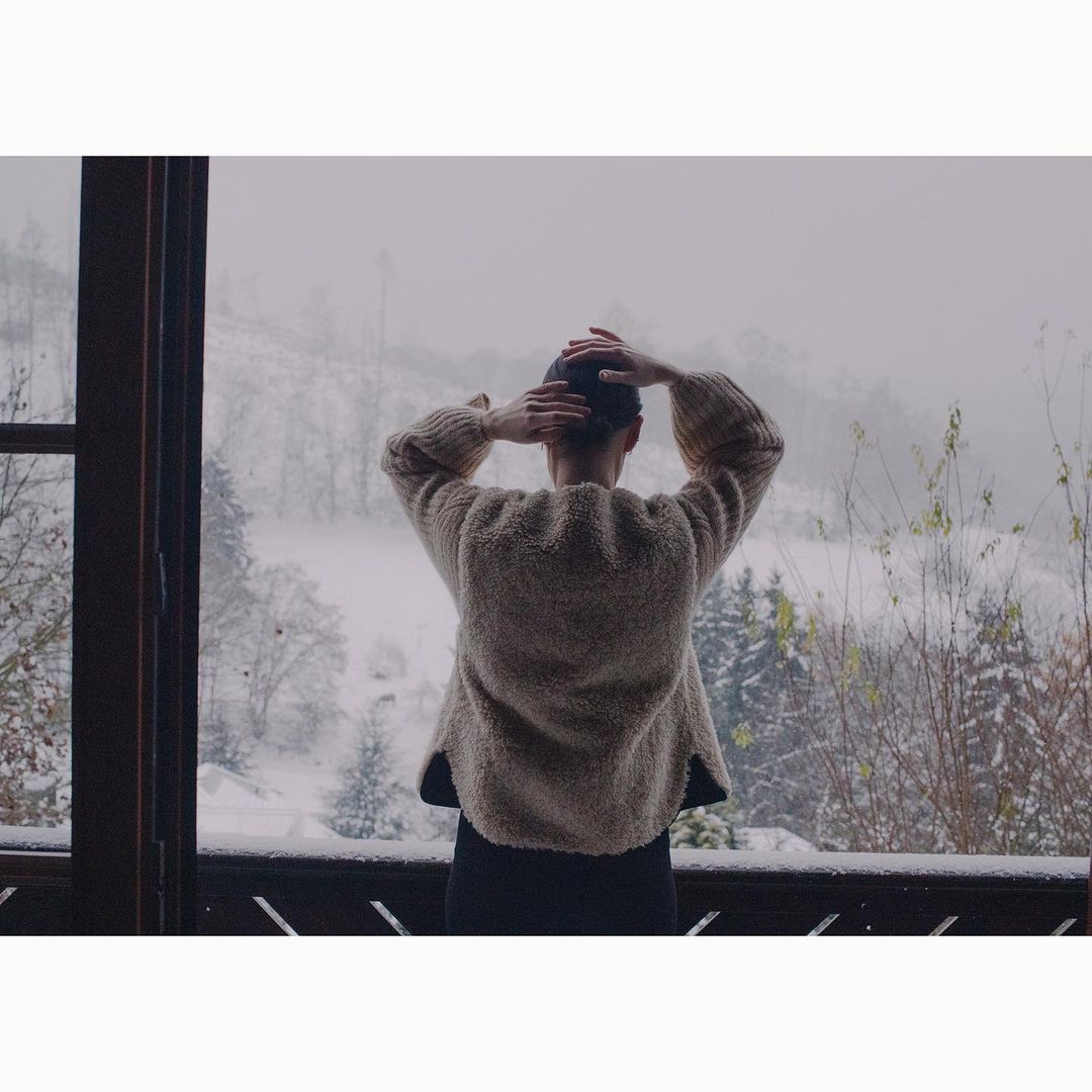 Adult female gazes over snow-covered mountain landscape from balcony of wooden house. Wearing a wool sweater, hands behind head.