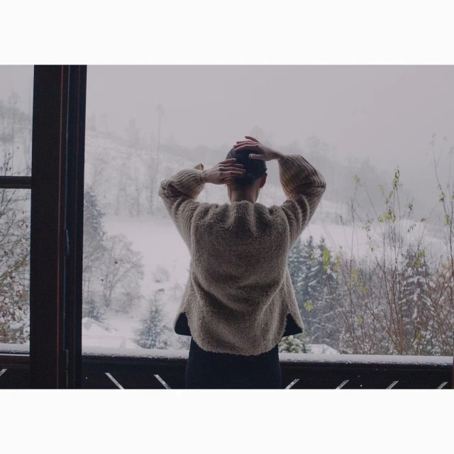 Adult female gazes over snow-covered mountain landscape from balcony of wooden house. Wearing a wool sweater, hands behind head.