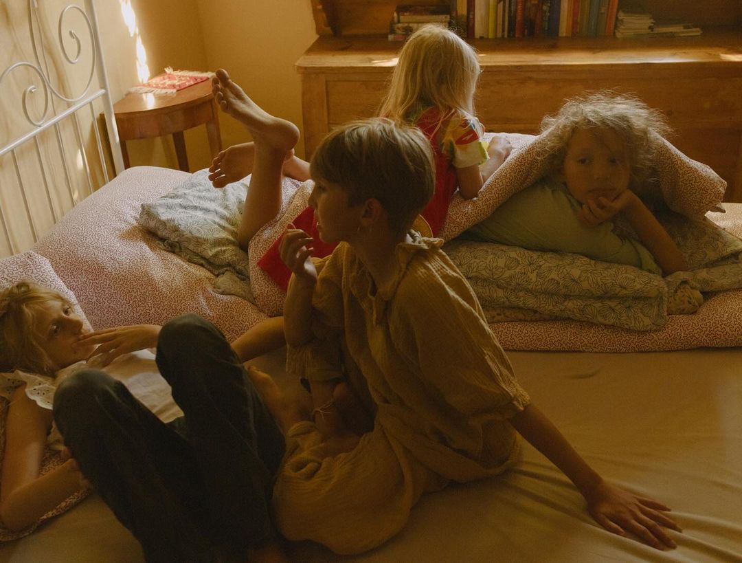 Four children relaxing on a bed with patterned sheets, blankets, and pillows in a warm-toned interior with wooden furniture and soft lighting.