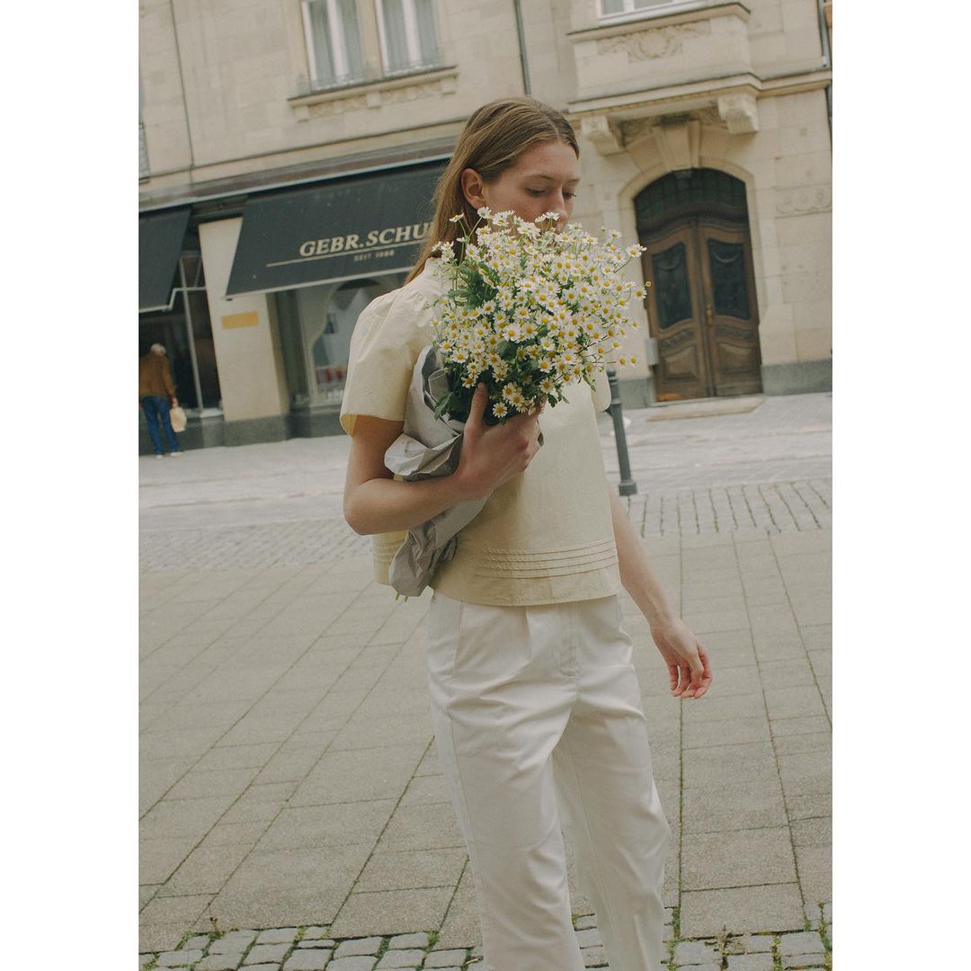 A blonde woman in an off-white blouse holds a bouquet of daisies on a city street with old buildings in the background on a sunny day.