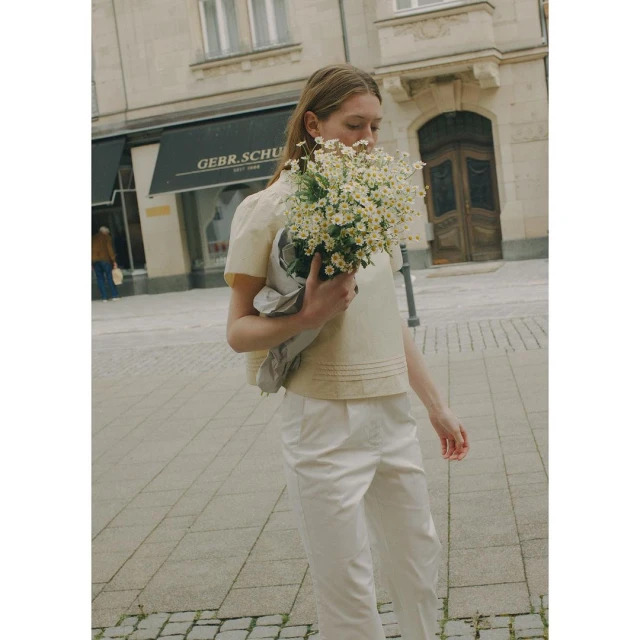A blonde woman in an off-white blouse holds a bouquet of daisies on a city street with old buildings in the background on a sunny day.