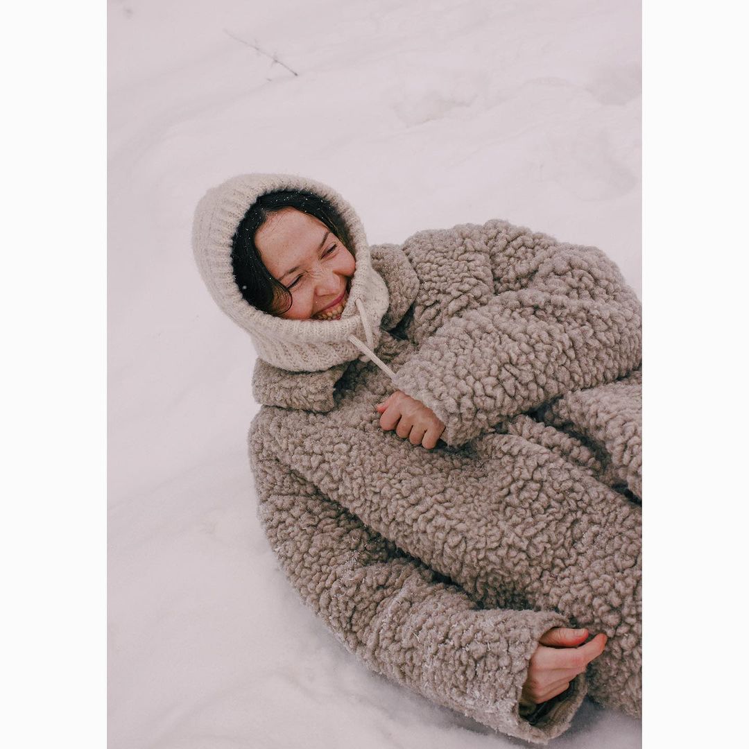 Woman in a hooded knit hat and a teddy-bear coat laying in the snow smiling broadly with a winter background.