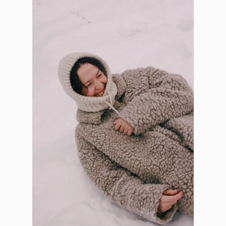 Woman in a hooded knit hat and a teddy-bear coat laying in the snow smiling broadly with a winter background.