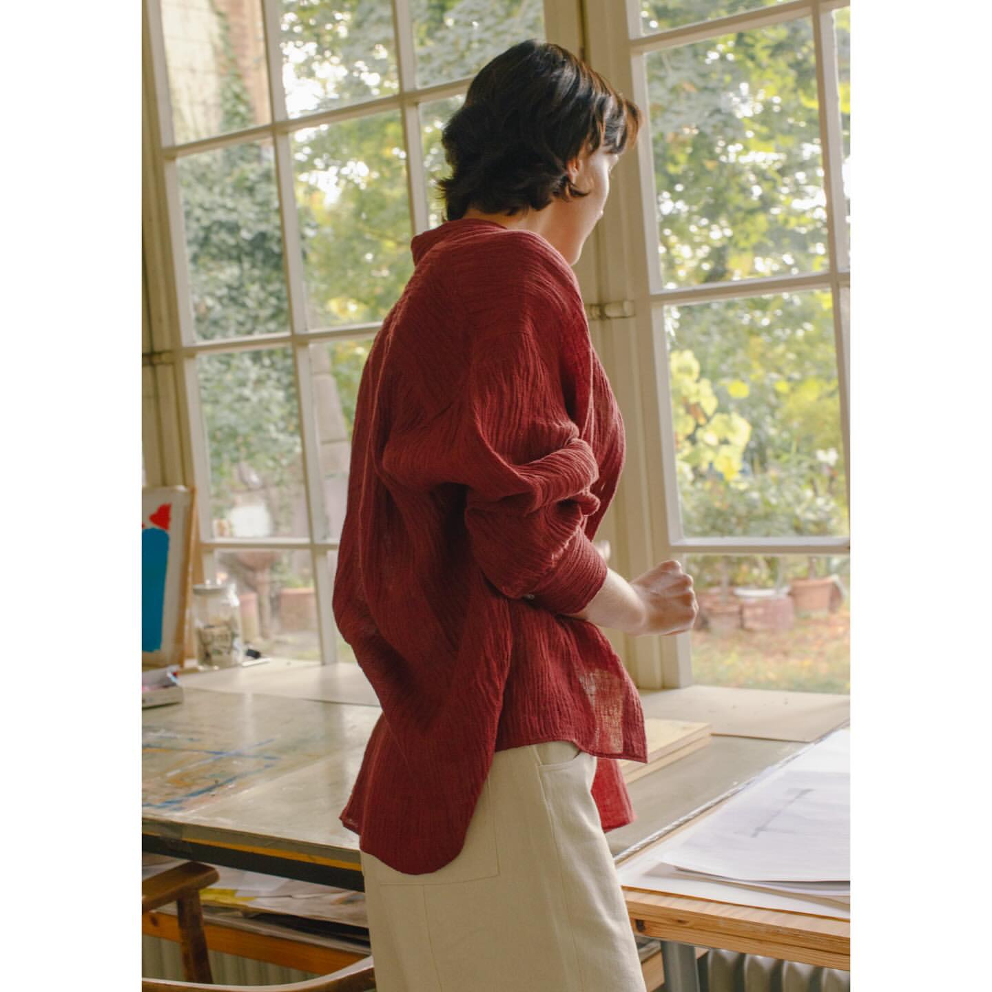 Woman in a red linen shirt stands near a window, looking out. Natural light illuminates the scene. Interior setting, neutral tones, and soft textures.