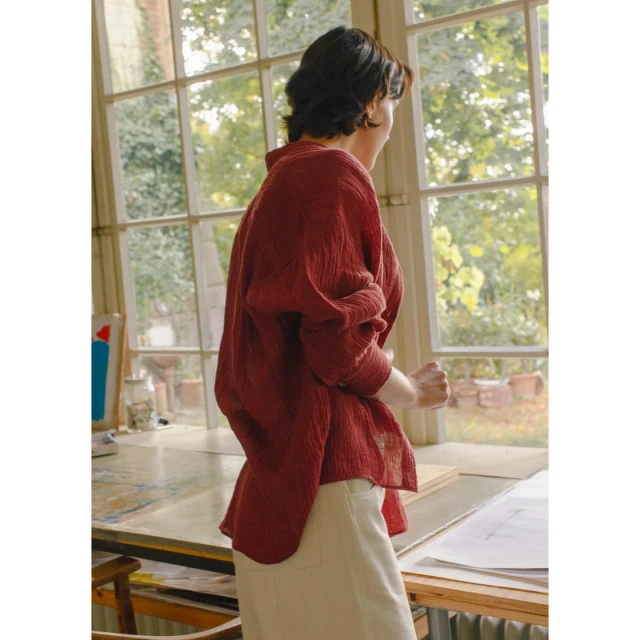 Woman in a red linen shirt stands near a window, looking out. Natural light illuminates the scene. Interior setting, neutral tones, and soft textures.