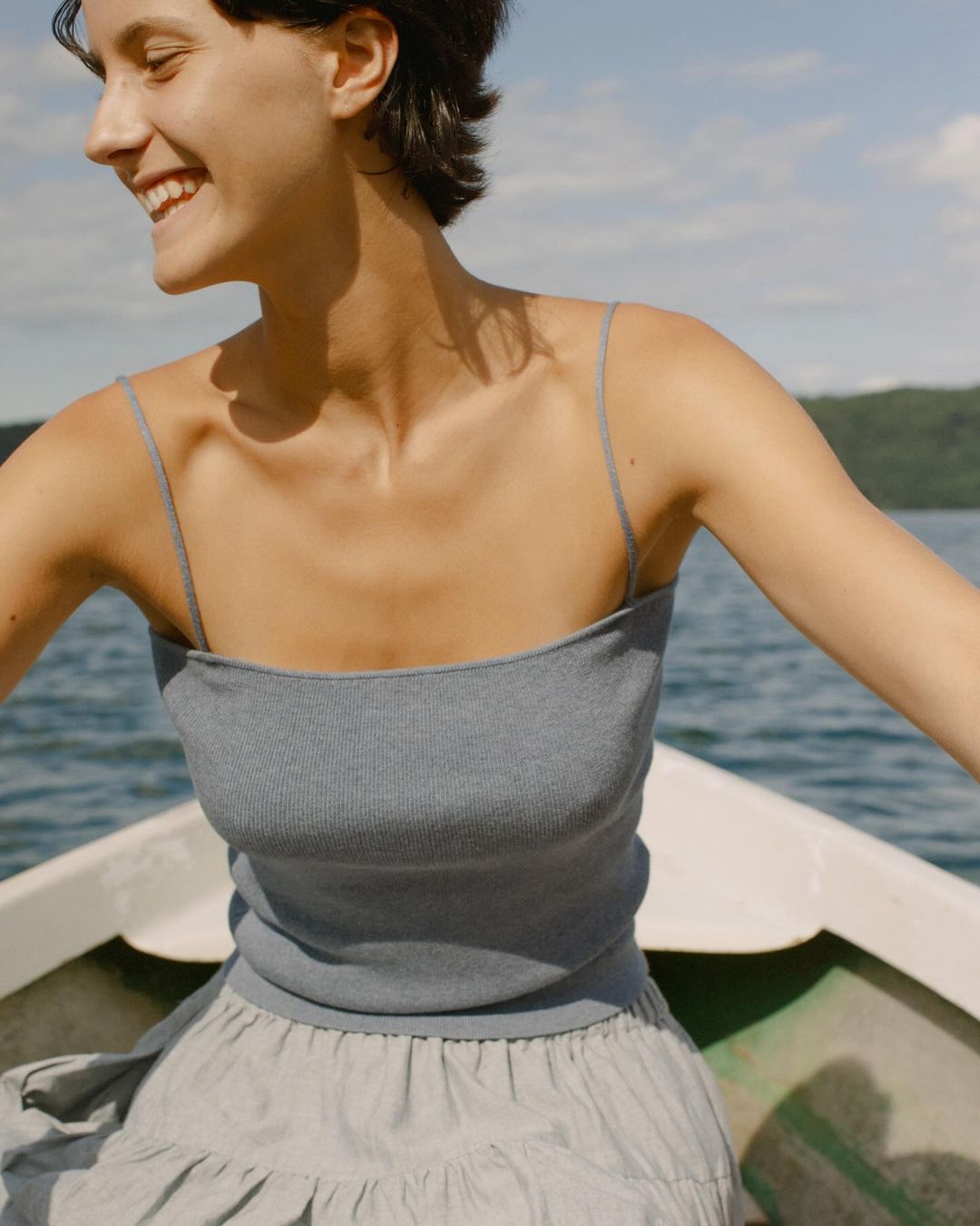 Young woman in boat smiles, wearing a blue top and skirt, with a lake and trees in the background on a sunny day. Lifestyle fashion.