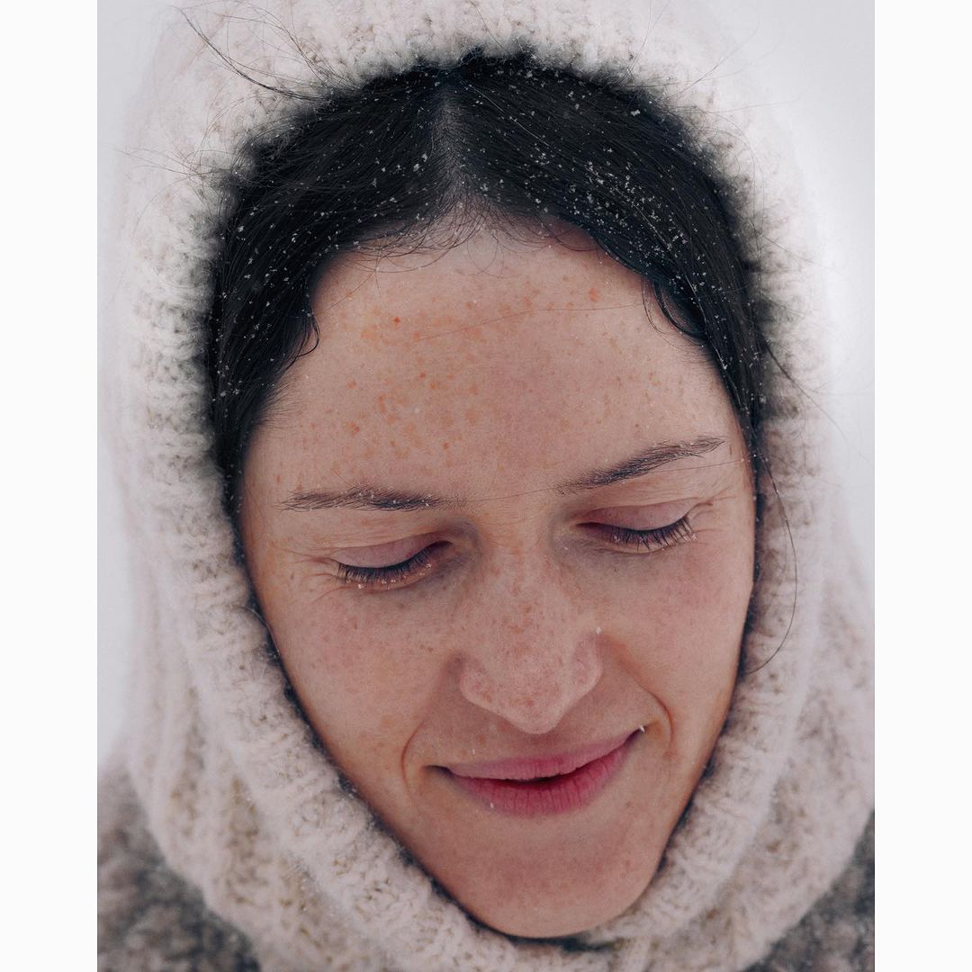 Close-up portrait of a smiling woman in a knitted hood, set against a bright background, showcasing her freckled skin and serene expression.