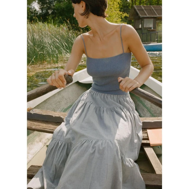Woman in a boat on a lake wearing a blue tank top and long gray dress, holding an oar, enjoying the summer season.