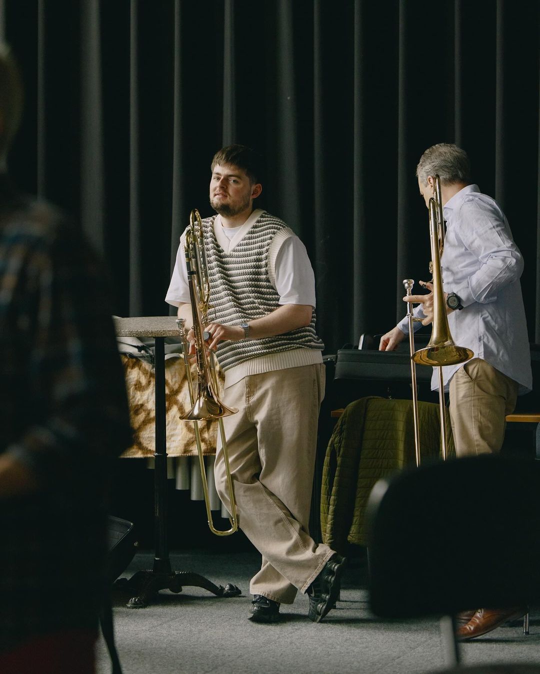 Two musicians with trombones in a music practice room, one leaning against a stand, the other near a keyboard and music equipment.