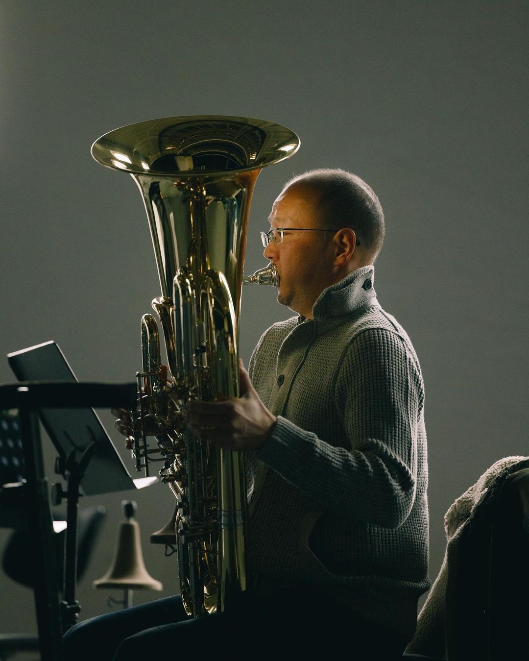 Man playing tuba indoors; a person wearing glasses and a sweater is playing a large brass instrument against a grey background.