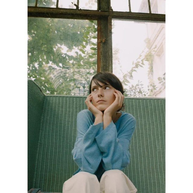 Woman with hands on face sits in front of a window overlooking a garden. She is wearing a blue top and light-colored pants.