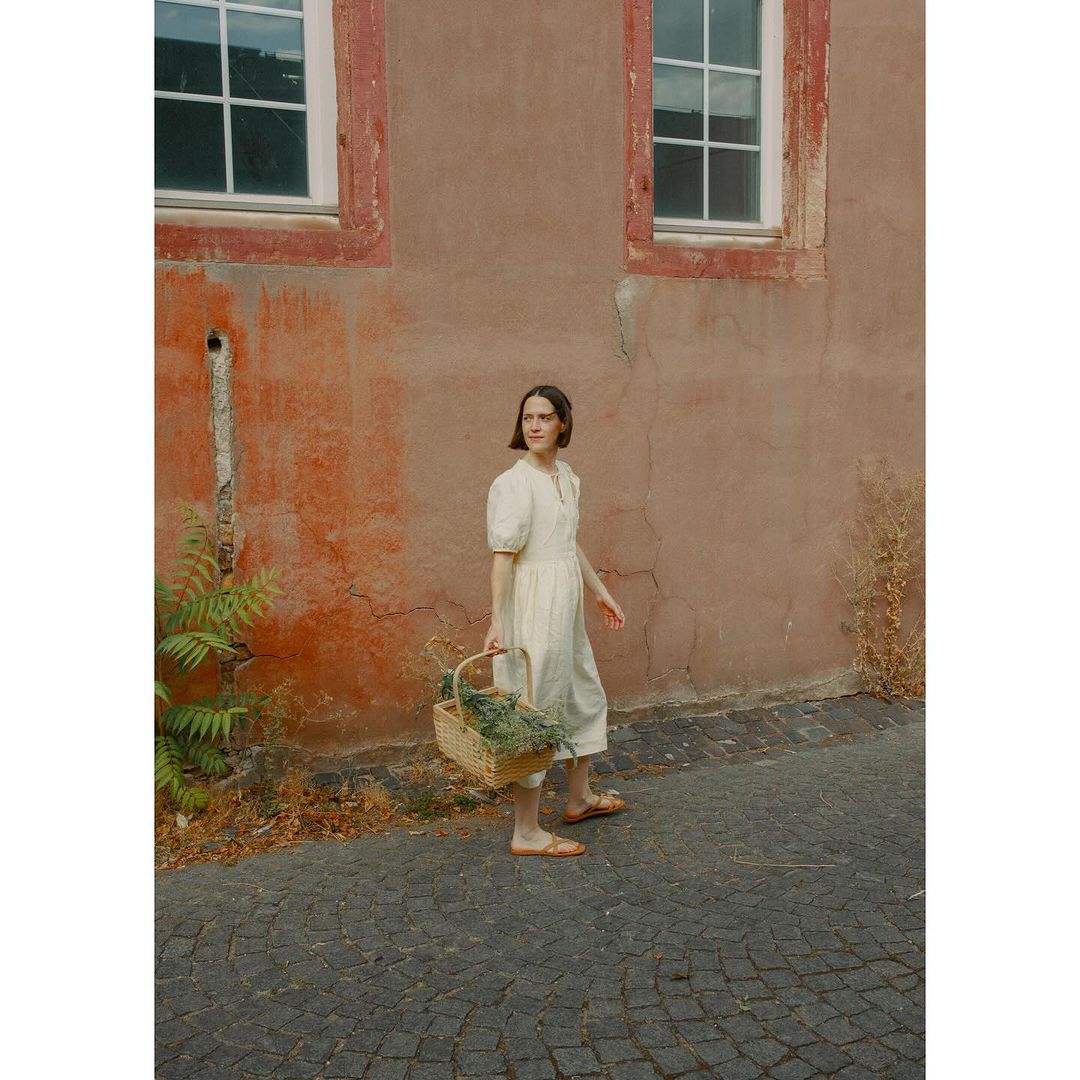 Adult female wearing a dress walks along a cobblestone street, carrying a woven basket in front of a peach colored building.