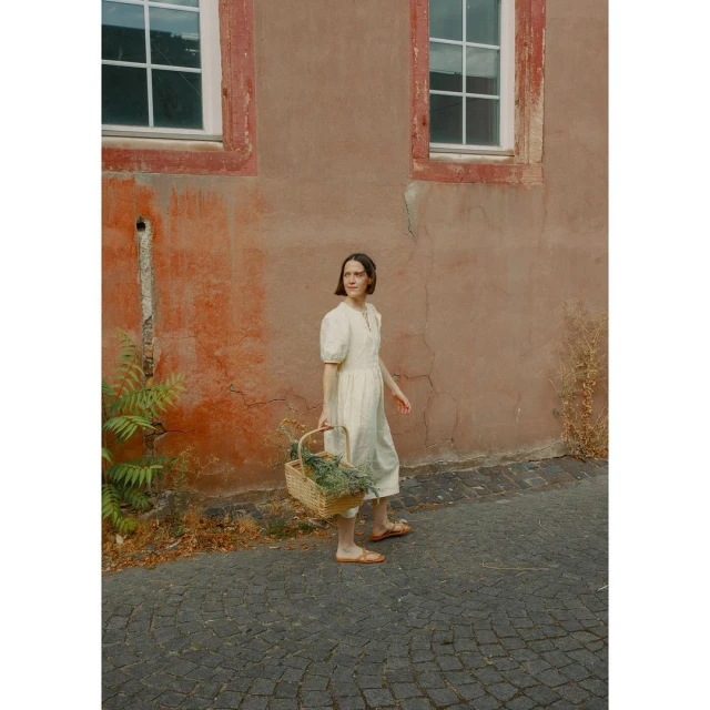 Adult female wearing a dress walks along a cobblestone street, carrying a woven basket in front of a peach colored building.