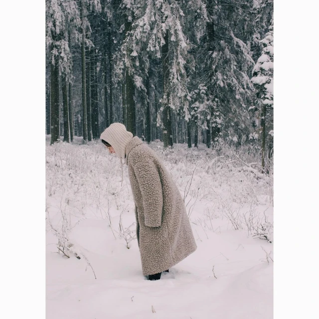 Adult female in wool coat and hat standing in snow. Forest with snow covered trees in the background. Soft, desaturated color palette.
