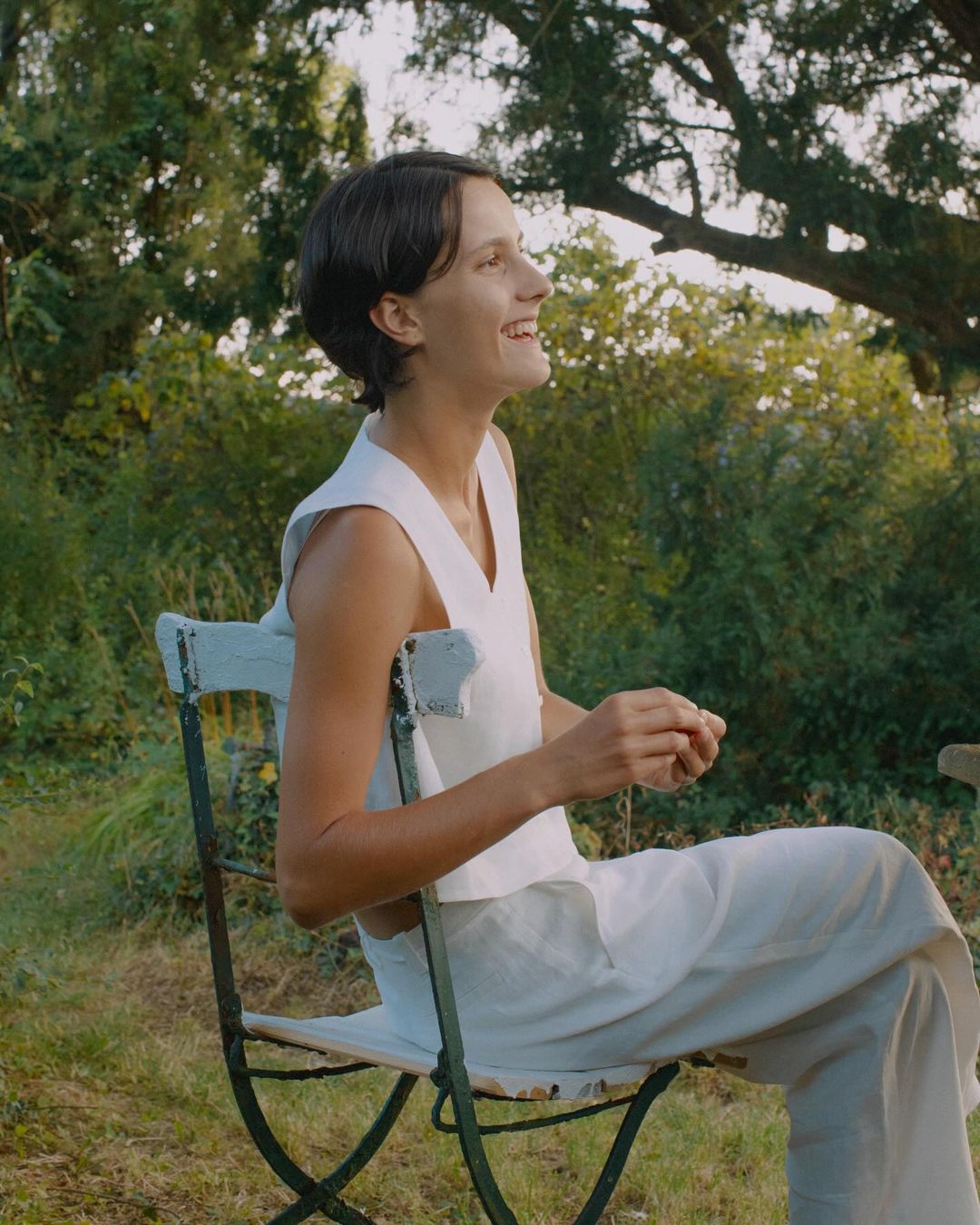 A smiling woman with short brown hair sits on a chair outdoors in a white top and pants with a leafy green background and dappled lighting.