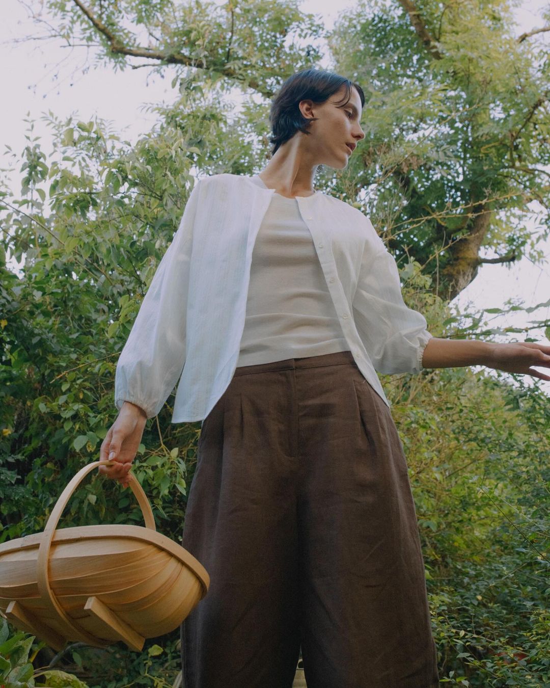 Woman in white blouse and brown pants holds a wooden basket outdoors in a wooded area, with green foliage in the background and natural lighting.